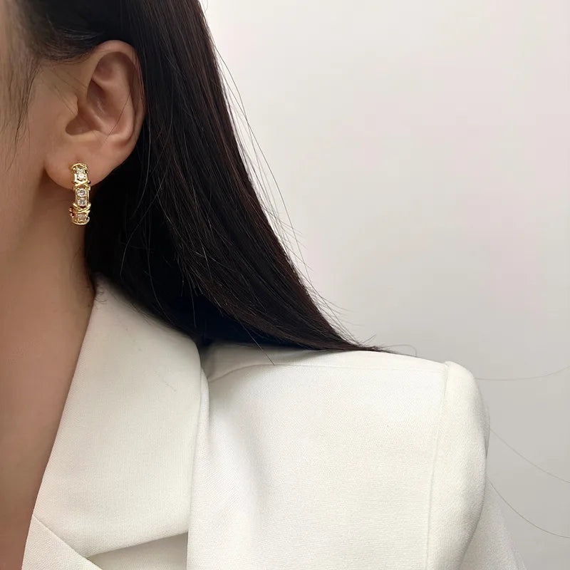 Créoles huggies dorées en acier, motif X pavé de zircons, portées sur l’oreille.