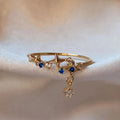 Bague ajustable en acier doré avec motif étoiles, pierres bleues et pendentif scintillant.