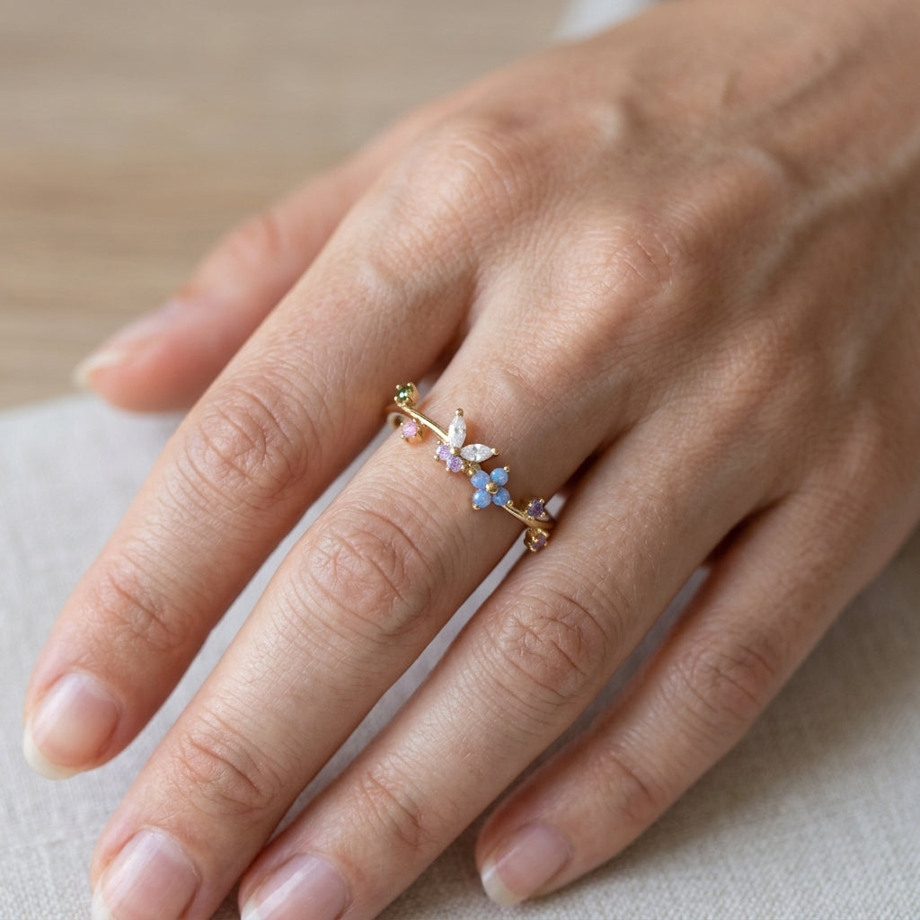 Bague ajustable dorée portée au doigt, motif jardin pastel avec fleurs bleues et papillon cristal – Jewelry by Kitana
