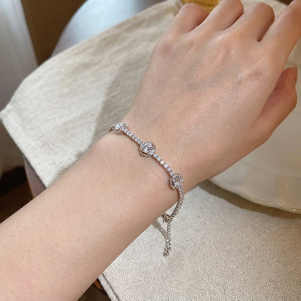 Bracelet rivière en acier inoxydable argenté porté au poignet, motifs camélia pavés de zircons scintillants, chaîne fine avec extension réglable, fond tissu beige

