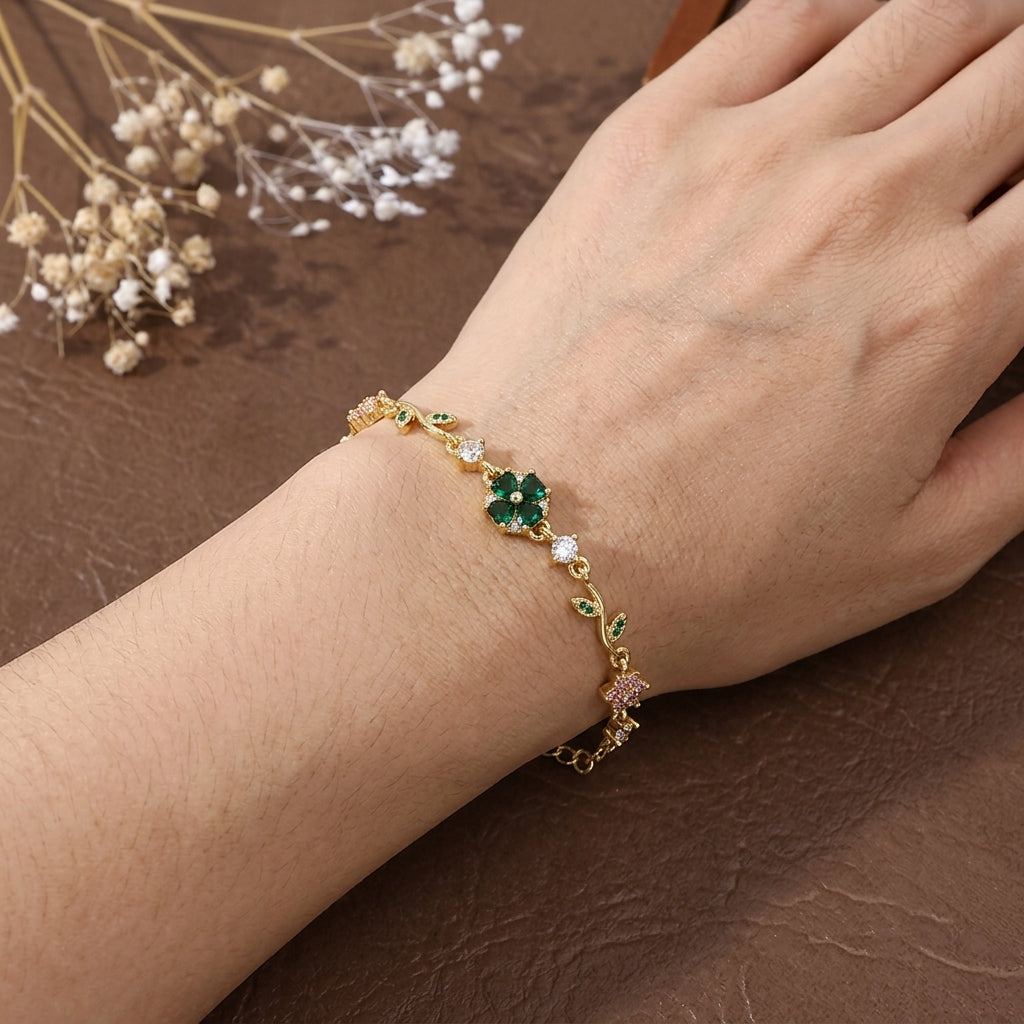 Bracelet plaqué or porté au poignet, motifs fleur trèfle en zircons émeraude et feuilles vertes, fond brun avec fleurs séchées blanches