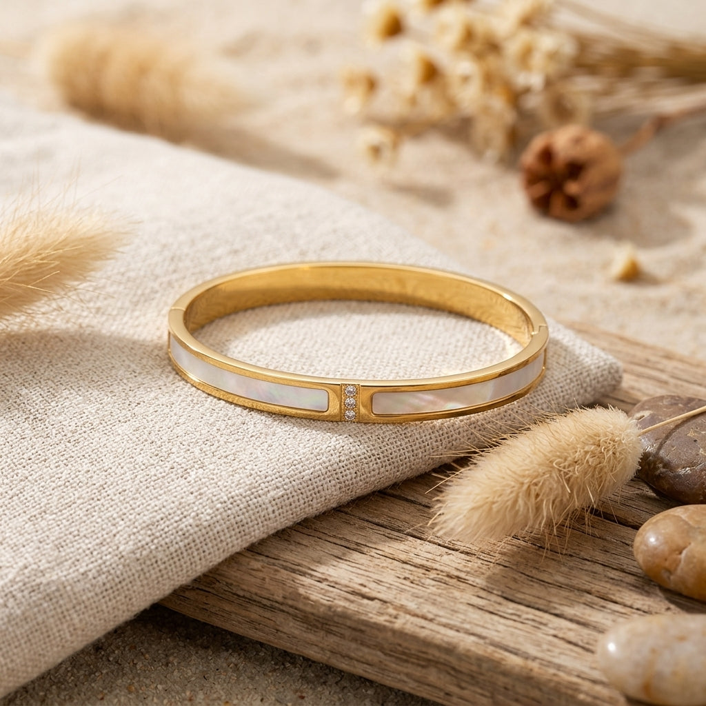 Bracelet jonc nacré doré avec strass posé sur un tissu clair et une surface en bois
Jewelry By Kitana 