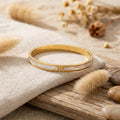 Bracelet jonc nacré doré avec strass posé sur un tissu clair et une surface en bois
Jewelry By Kitana 